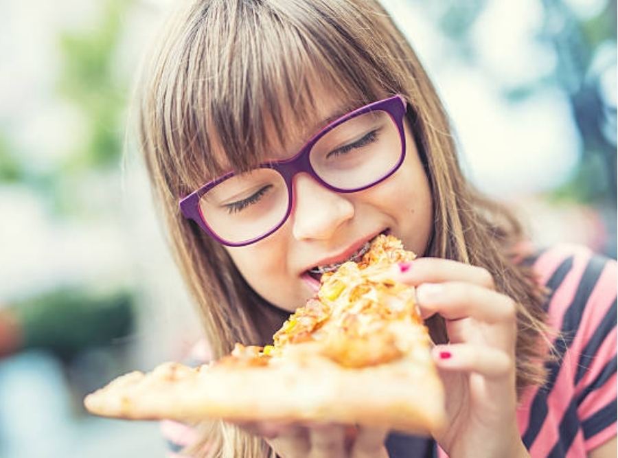 Child wearing braces holding a slice of pizza outdoors, illustrating kids enjoying food while undergoing orthodontic treatment.
