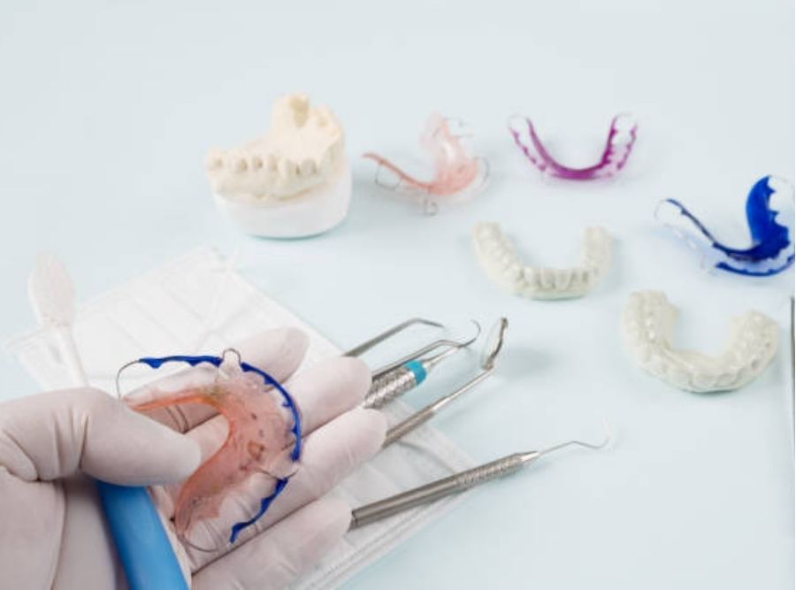 Close-up of a gloved hand holding a colorful orthodontic retainer beside dental tools and plaster teeth models, displaying different removable orthodontic appliances on a light background.