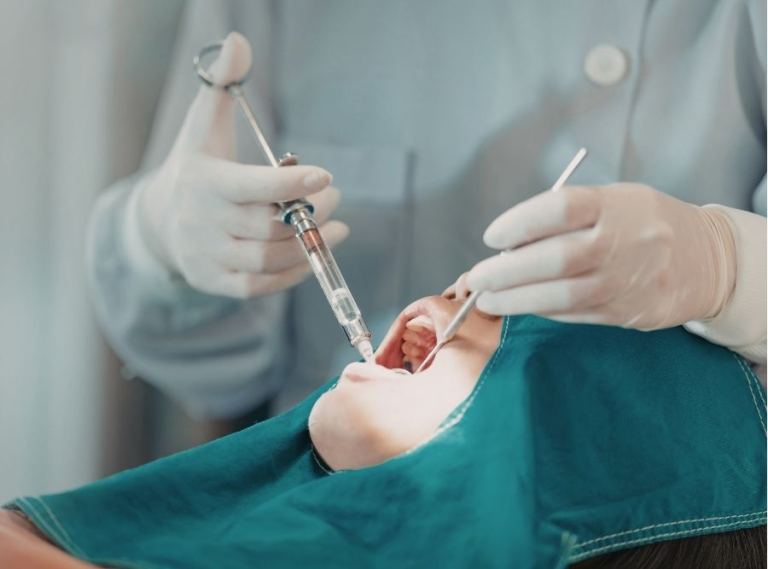 Dentist administering local anesthesia injection for orthodontic or dental treatment, close-up of gloved hands holding syringe and dental mirror while patient lies on chair covered with protective drape.