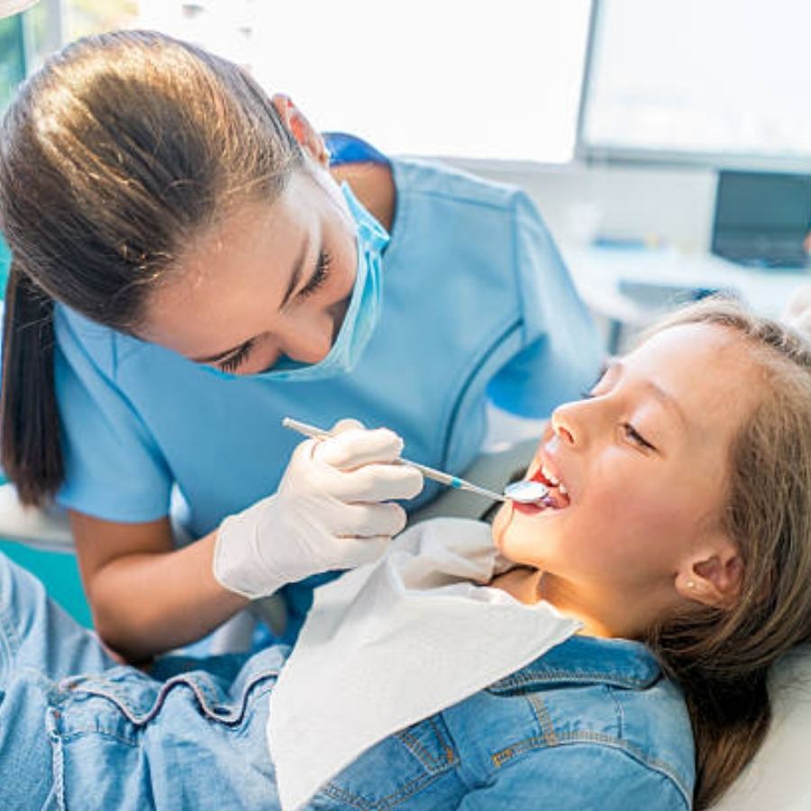 Female dentist in blue scrubs and a face mask performing a dental checkup on a young girl using a dental mirror